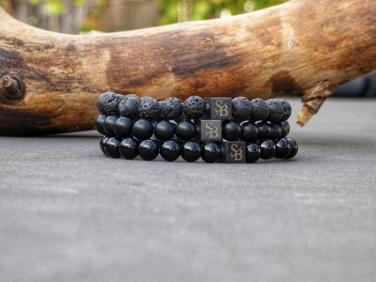 Drie zwarte natuursteen armbanden naast elkaar op beton: poreuze lavasteen, matte obsidiaan en glanzende obsidiaan