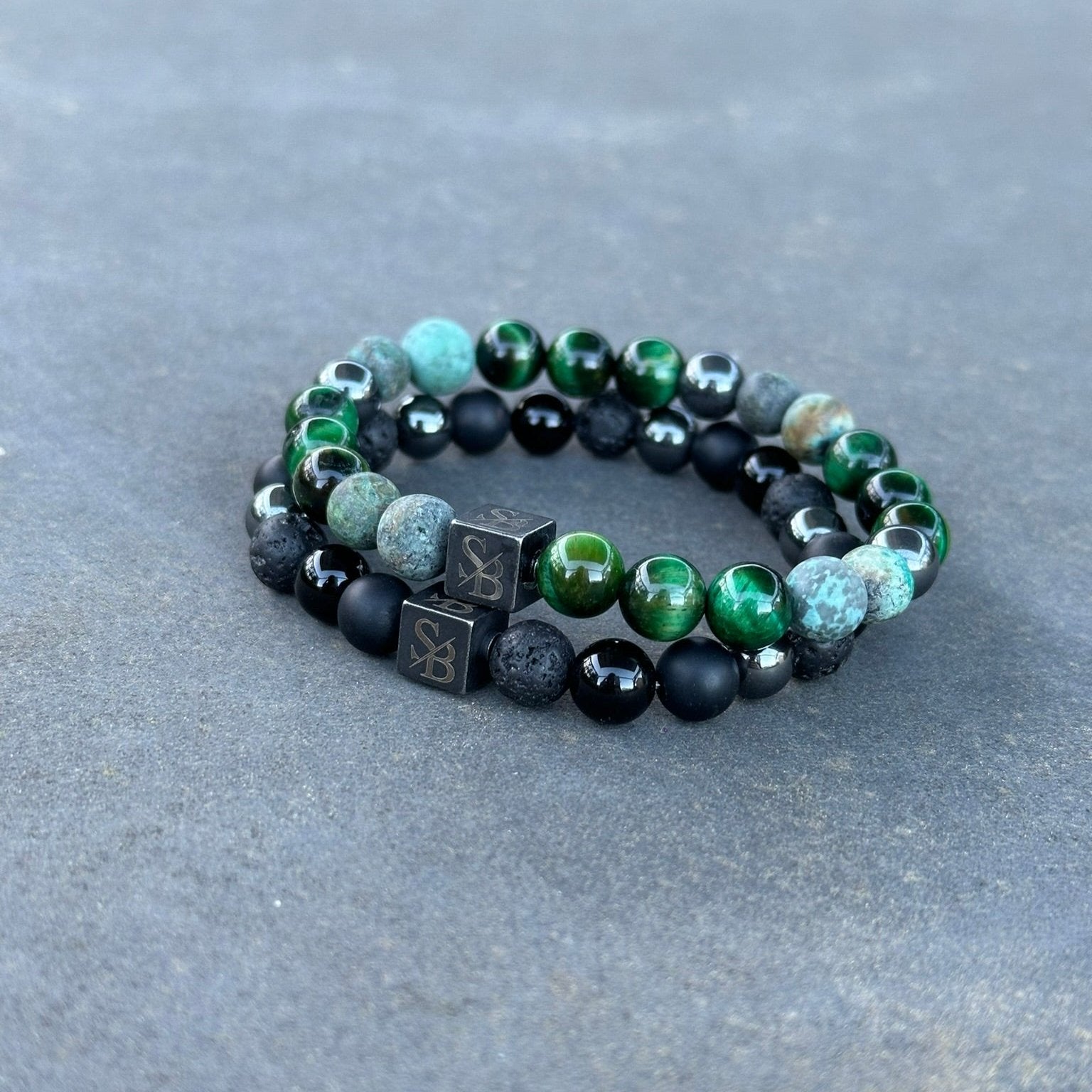 Een paar Swamp | Mixed Stones (8mm) kralenarmbanden van Stoney Bracelets worden getoond tegen een grijze achtergrond. De armbanden hebben een combinatie van groene, zwarte en steenkleurige kralen, waaronder kubusvormige kralen met symbolen erop. Gemaakt met natuursteen en edelsteen kralen, zijn de armbanden opgerold en liggen ze naast elkaar.