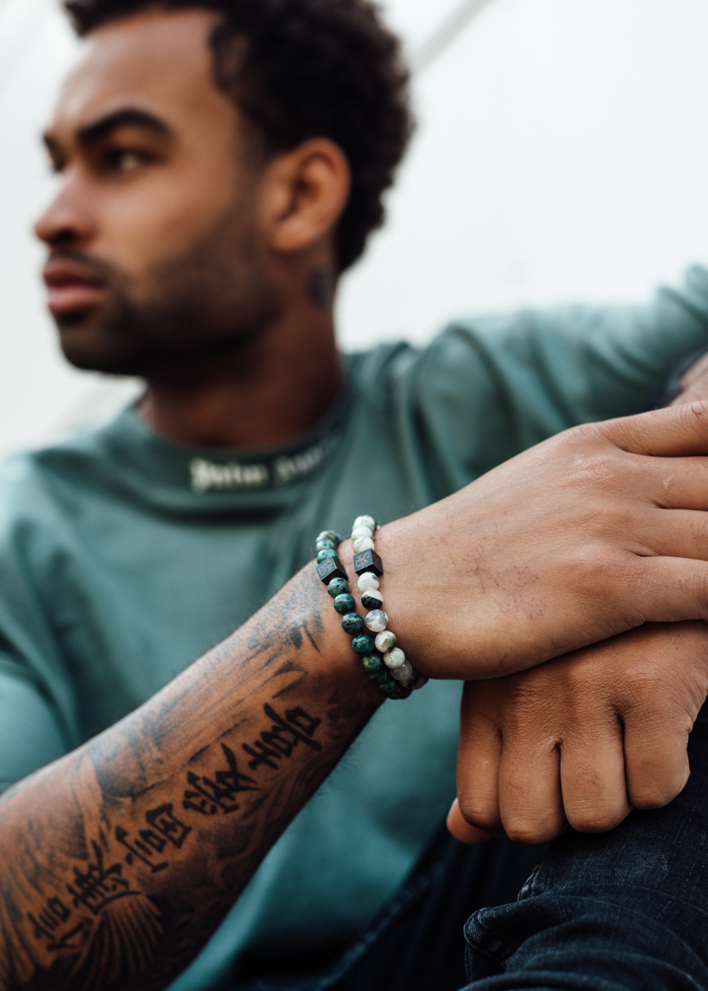 Een persoon met kort krullend haar, gekleed in een groen shirt, zit en kijkt opzij. Hun arm rust op hun knie, met tatoeages en kralenarmbanden van natuursteen. De achtergrond is onscherp, waardoor het individu en hun unieke accessoires worden benadrukt.