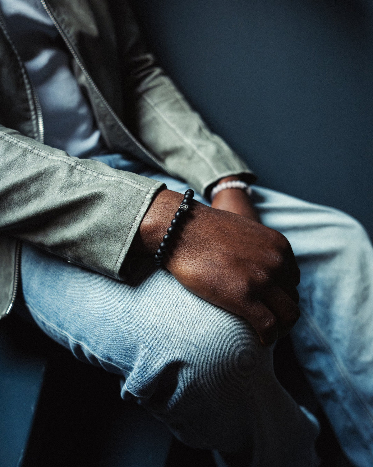 Een getatoeëerde arm rust op een betonnen oppervlak. De persoon draagt twee kralenarmbanden, een witte en een groene en zwarte. Een gedeeltelijke weergave van een wit shirt met een logo is zichtbaar.