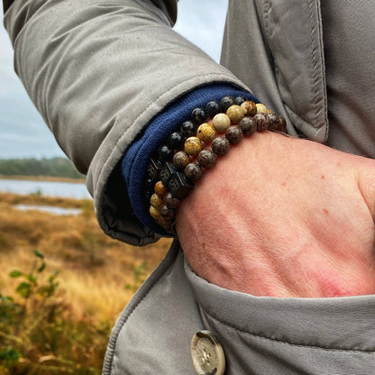 Een close-up van de pols van een persoon die de Triple Brown Set (8mm) kralenarmbanden van Stoney Bracelets draagt, met verschillende kleuren waaronder bruin en beige. De persoon is gekleed in een grijs jasje over een blauwe mouw, staand in een buitenomgeving met grasachtig terrein en een watermassa op de achtergrond.