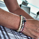 Een close-up van een arm versierd met Stoney Bracelets' "Matte Zebra Stones (6mm)" armbandset rustend op een geruit stoffen oppervlak. Tussen de kleurrijke strengen met witte, grijze en roze kralen met kleine zwarte blokjes zitten hints van edelsteen. De wazige achtergrond onthult een onscherpe buitenomgeving met een stoel en tafel.