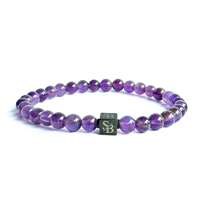 Een armband van Stoney Bracelets gemaakt van 6mm ronde, gepolijste Amethist Stones kralen die aan elkaar geregen zijn in een cirkel. De armband heeft een vierkante metalen kraal met de gegraveerde initialen "SB" in het midden, tegen een effen witte achtergrond.