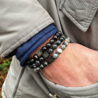 Close-up van een persoon die meerdere kralenarmbanden om zijn pols draagt. De armbanden, van Stoney Bracelets' Triple Gray Set (8mm), zijn voorzien van kralen in zwart, wit en grijs, sommige met patronen die lijken op natuursteen zoals marmer of graniet. De persoon is gekleed in een beige jasje en een donkerblauwe mouw piept onder de mouw van het jasje vandaan.