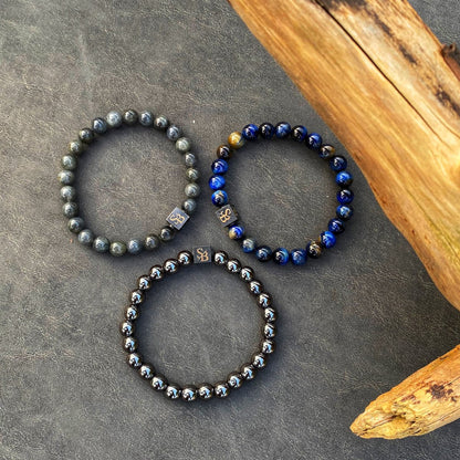 Drie armbanden uit de Stoney Bracelets Triple Blue Set (8mm) worden op een donker getextureerd oppervlak naast een stuk drijfhout getoond. Elke armband heeft edelsteen kralen in verschillende kleuren: grijs, diepblauw met gouden accenten en metallic zwart. Kleine metalen bedelkralen met symbolen zijn tussen de kralen verspreid.