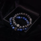 De Lapis Lazuli Set (8mm) van Stoney Bracelets wordt gepresenteerd in een strak zwart doosje, met twee kralenarmbanden. Eén armband benadrukt donkergrijze natuursteen kralen, terwijl de andere armband afwisselt tussen donkergrijze en blauwe edelsteen kralen. Elke armband is versierd met een klein vierkant bedeltje gegraveerd met het logo van het merk. De achtergrond presenteert een rijke, gestructureerde stof.