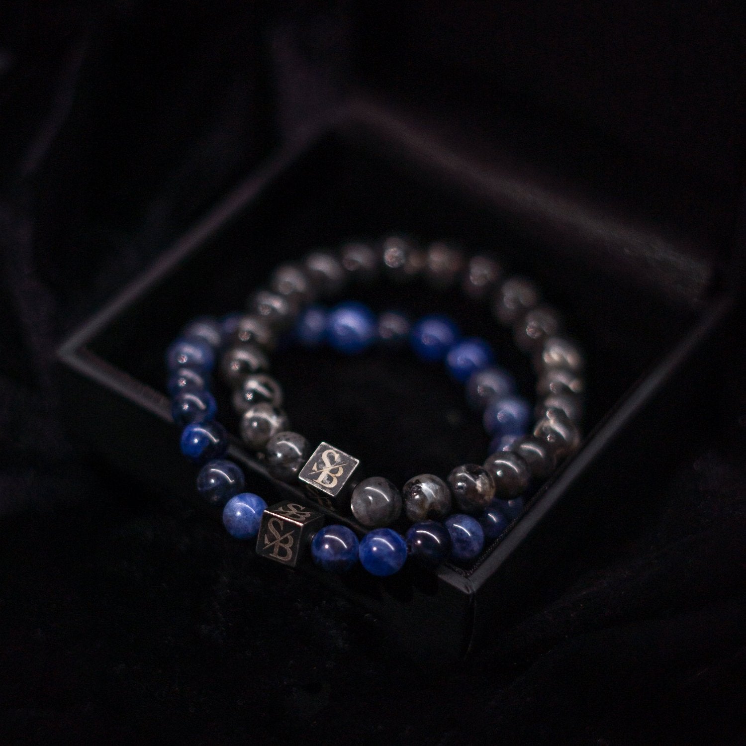 Een foto van twee kralenarmbanden uit de Moon Stones (8mm) collectie van Stoney Bracelets in een zwart doosje. De armbanden bestaan uit donkere en blauwe natuurstenen kralen, elk met een klein vierkant metalen bedeltje met de initialen "SY" erin gegraveerd. Het doosje is gevoerd met zwarte stof.