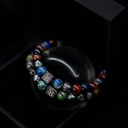 Close-up van twee Stoney Bracelets' Greek Set (8mm) armbanden op een zwarte achtergrond. De armbanden zijn voorzien van kleurrijke kralen in tinten blauw, groen, rood en zilver, met vierkante metalen accenten met een gegraveerd symbool.