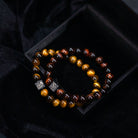 Twee kralenarmbanden van Stoney Bracelets, met gepolijste Red Tiger Eye Stones (8 mm) in tinten bruin, goud en zwart, zijn naast elkaar geplaatst op een donker, fluwelen doosje. Elke armband is versierd met een enkele vierkante metalen kraal gegraveerd met een decoratief patroon. Het doosje rust op een zwarte stoffen achtergrond.