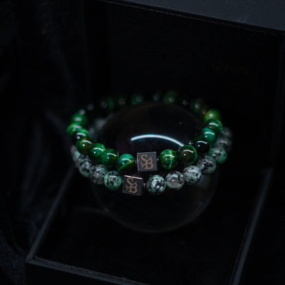 Een close-up van twee kralenarmbanden van Stoney Bracelets. De ene is versierd met groene tijgeroogstenen (8 mm), terwijl de andere is versierd met zwarte en witte gespikkelde stenen. Elke armband heeft een klein vierkant bedeltje met de initialen "SB" gegraveerd en wordt weergegeven op een strak zwart oppervlak.