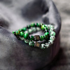 Op een grijze stof zijn twee armbanden uit de "Nature Set (8mm)" van Stoney Bracelets te zien. De ene heeft groene kralen in donker- en lichtgroene patronen, terwijl de andere een gevlekt ontwerp van witte en groene kralen toont. Beide armbanden bevatten een klein zwart vierkant bedeltje versierd met goudkleurige initialen "SB.