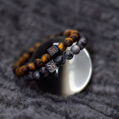 Twee kralen armbanden uit de Hunter Set (8mm) van Stoney Bracelets, één met tijgeroog kralen en de andere met grijs gemarmerde natuursteen, zijn gedrapeerd over een reflecterende glazen bol. Beide armbanden zijn voorzien van vierkante zwarte kralen met de initialen "SB" in metallic letters. De achtergrond is een zachte, donkergrijze stof.