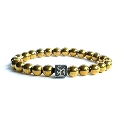 Een goudkleurige bolvormige kralenarmband van Stoney Bracelets, met 8 mm gouden Hematiet-stenen, wordt op een witte achtergrond weergegeven. Het bevat een vierkante bedel met de letters "SB" in het midden gegraveerd.