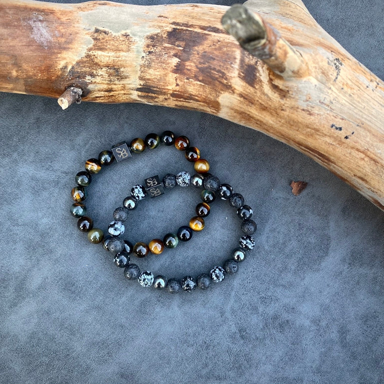 Twee kralenarmbanden liggen op een grijs oppervlak met daarboven een stuk drijfhout. Eén armband, van Stoney Bracelets, heeft Multicolor Tiger Eye Stones (8 mm) in zwart en wit gemarmerde kralen, terwijl de andere zowel donkere als metaalachtige kralen toont, versterkt met verbluffende natuursteenaccenten.