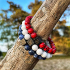 De kralenarmbanden "Red Vintage Stones (8mm)" van Stoney Bracelets, met een mix van rode, witte en blauwe ronde kralen naast vierkante metalen kralen met symbolen erin gegraveerd, zijn kunstig gedrapeerd rond een boomtak. Sommige kralen zijn gemaakt van natuursteen. De achtergrond toont zacht vervaagde bomen en lucht.