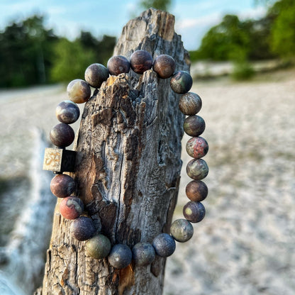 Een kralenarmband met 8mm Flower Jasper Stones van Stoney Bracelets is om een verweerd stuk hout op het strand gewikkeld. De achtergrond toont zanderige grond en enigszins onscherpe bomen.