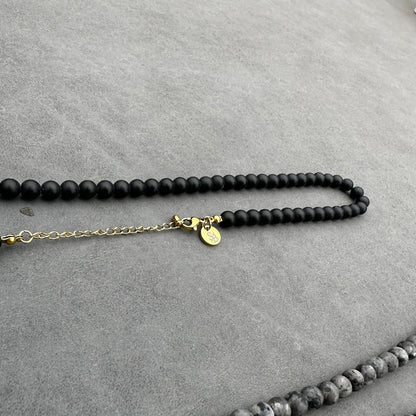 De Matte Obsidian Necklace (6 mm) van Stoney Bracelets heeft matte zwarte kralen op een grijs oppervlak, samen met een gouden verlengstuk en een ronde gouden bedel, die kracht en bescherming symboliseren.