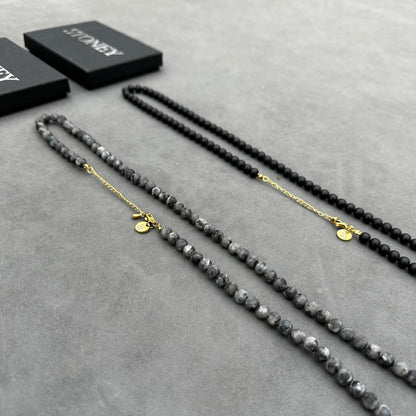 Op een grijs oppervlak heeft de Matte Moon Necklace van Stoney Bracelets elegante grijze kralen en straalt kalmte uit. Ernaast zit een andere ketting met opvallende zwarte kralen; beide hebben gouden sluitingen en ronde labels, allemaal gedeeltelijk genesteld in zwarte dozen.