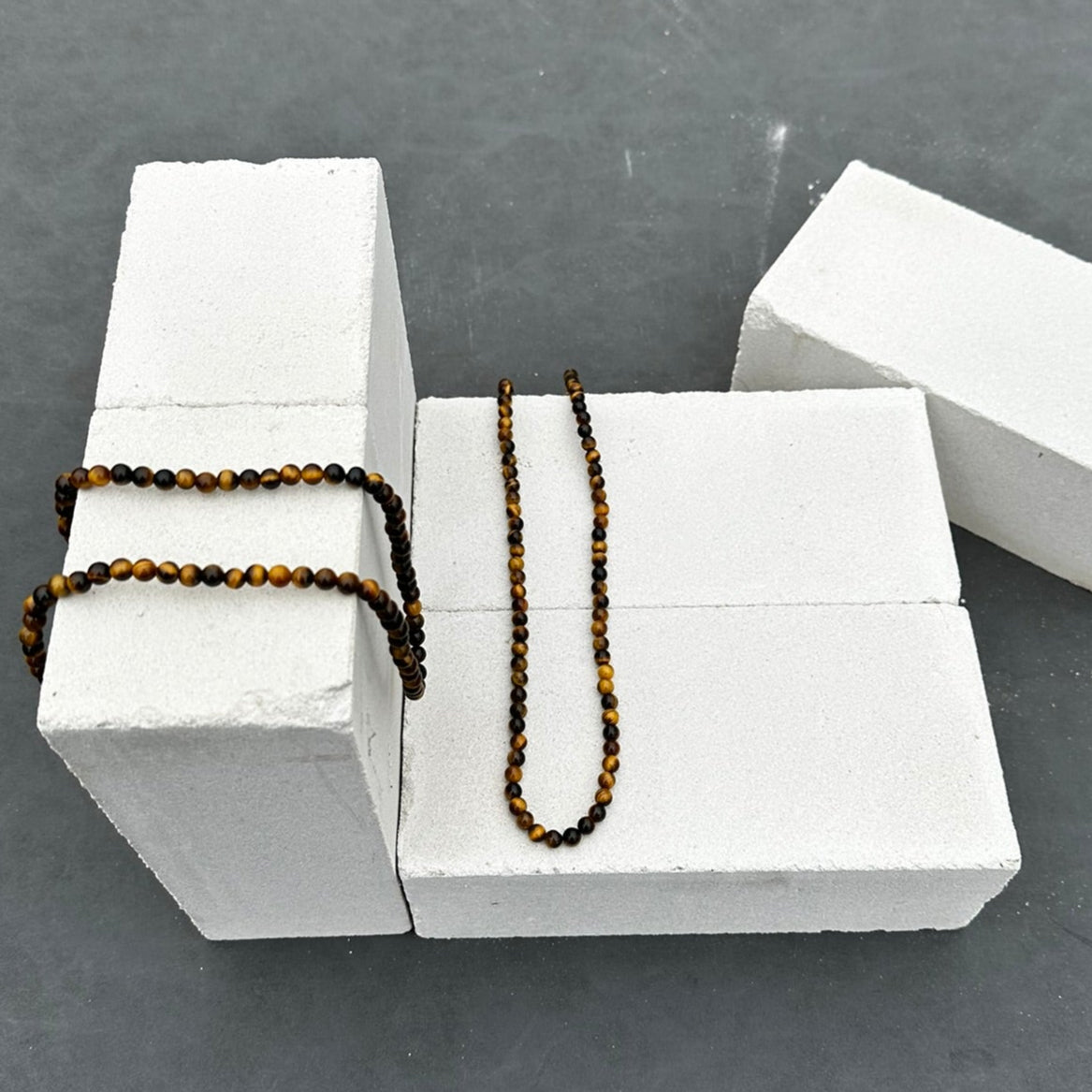 De Stoney Bracelets' Golden Gaze Necklace (6mm) is afgebeeld op witte stenen en is gemaakt van gepolijste Tiger Eye-kralen. Hij valt elegant over de rand met een effen grijze achtergrond die zijn aantrekkingskracht versterkt.