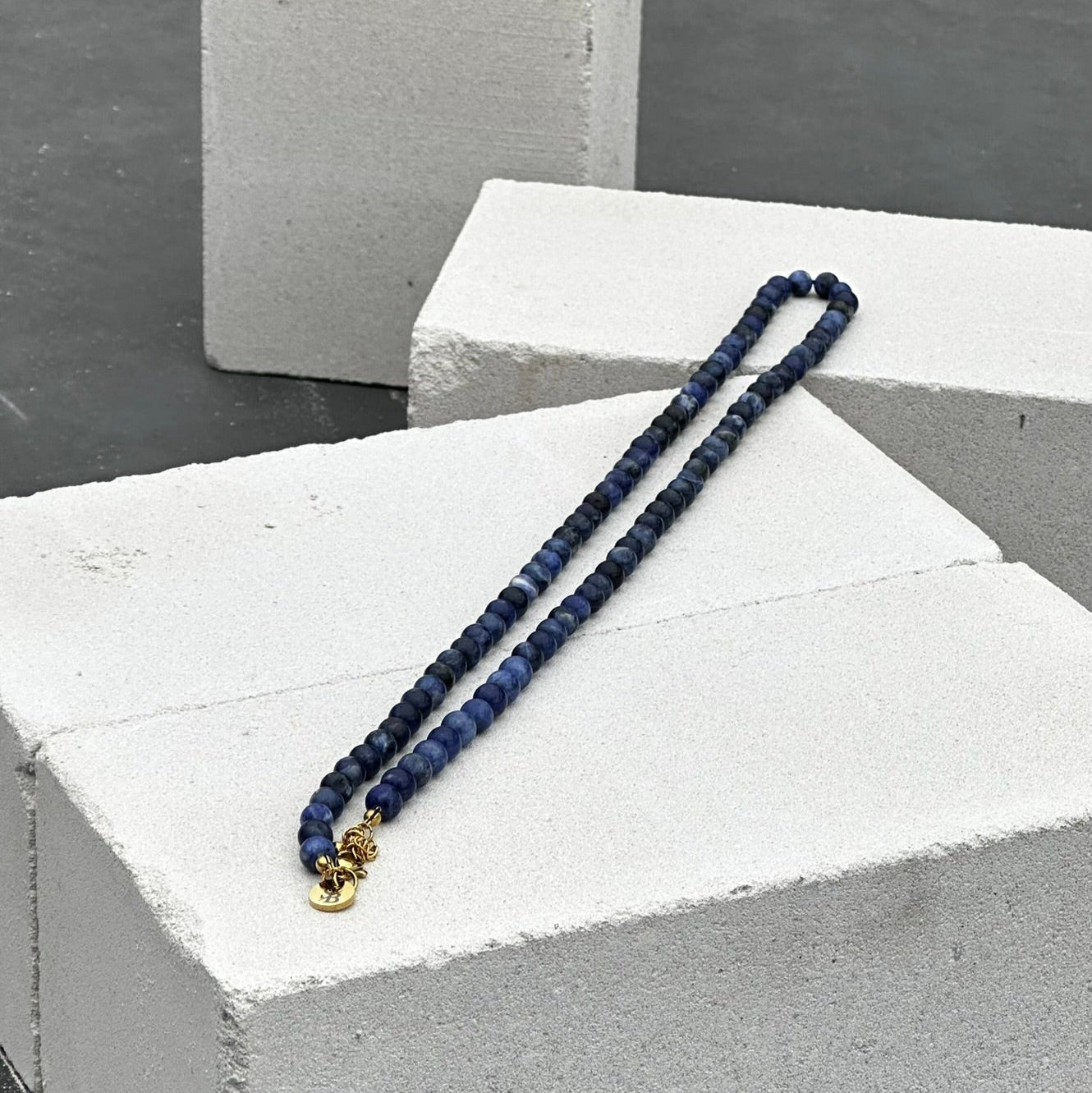 De Royal Lapis Necklace (6mm) van Stoney Bracelets, met blauwe kralen en een klein gouden bedeltje, rust elegant op witte sintelblokken. De zachte curve tegen een donkere achtergrond creëert een verbluffend statement piece.