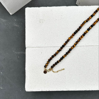 De Golden Gaze Necklace (6 mm) van Stoney Bracelets heeft een witte achtergrond met structuur en een steenachtig grijs materiaal. De ketting is voorzien van gouden tijgeroogkralen en een gouden ketting en sluiting.