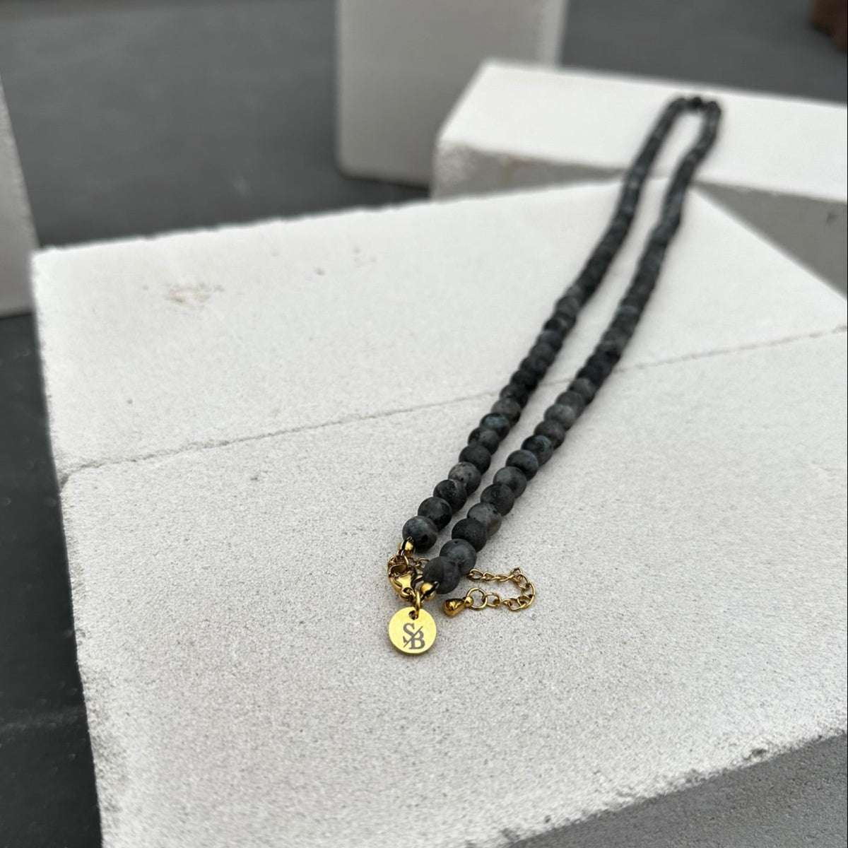 De Stoney Bracelets Matte Moon Necklace (6 mm) heeft een zwart kralenontwerp met een klein gouden kettinkje en een ronde hanger die emotionele balans symboliseert. De ketting rust op een wit oppervlak met structuur en is versierd met soortgelijke serene witte blokken.