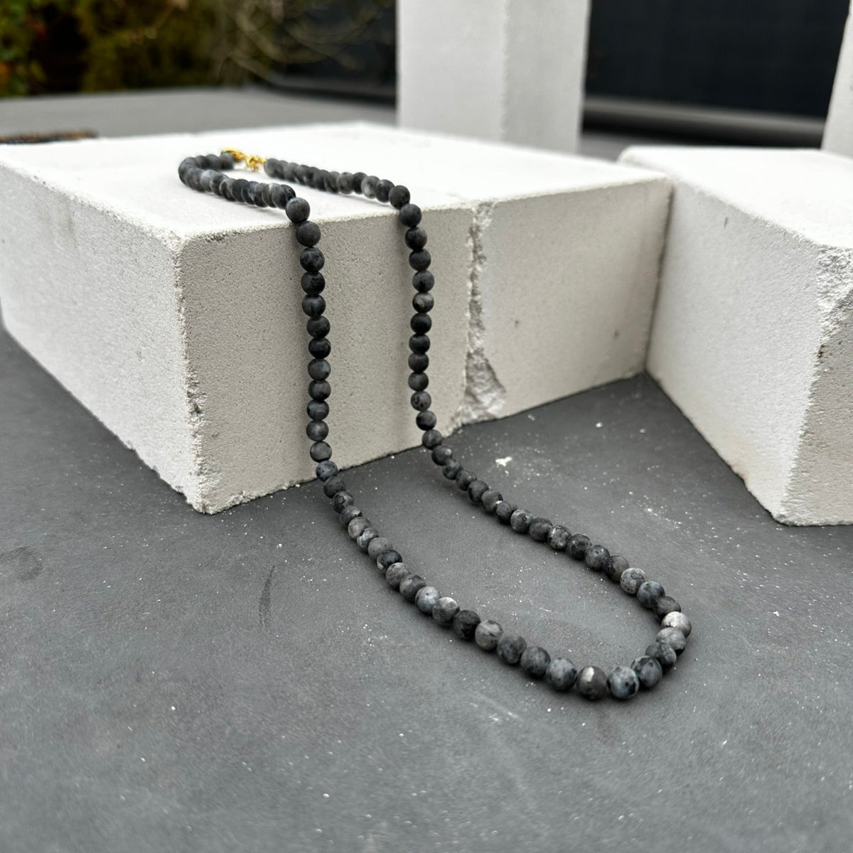 De Matte Moon Necklace (6 mm) van Stoney Bracelets, met donkere ronde kralen en een gouden sluiting, rust elegant op ruwe witte betonblokken buiten en straalt emotionele balans uit.