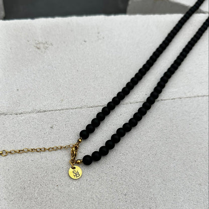 De Matte Obsidian Necklace (6mm) van Stoney Bracelets heeft een gouden sluiting en ketting die rust op een gestructureerd oppervlak. Het toont elegant een kleine ronde gouden bedel met een gegraveerd symbool voor kracht en bescherming, wat elegantie en sterkte belichaamt.