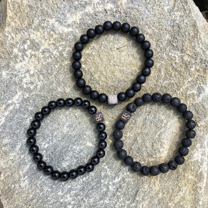 De Triple Black Set (8mm) van Stoney Bracelets is gerangschikt op een grijs natuursteen oppervlak. Elke armband toont een verscheidenheid aan zwarte kralentexturen en ze zijn allemaal versierd met een kleine, kubusvormige metalen bedel gegraveerd met ingewikkelde symbolen.