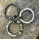 Drie edelsteen kralen armbanden uit Stoney Bracelets' Marble Stones collectie (8mm) worden getoond op een stenen oppervlak. De bovenste linker armband heeft donkergrijze gemarmerde kralen, de bovenste rechter armband heeft uniforme witte kralen en de onderste armband toont een mix van witte, groene en grijze gemarmerde kralen. Elke armband is versierd met een kleine zwarte vierkante bedel gegraveerd met de initialen "SB.