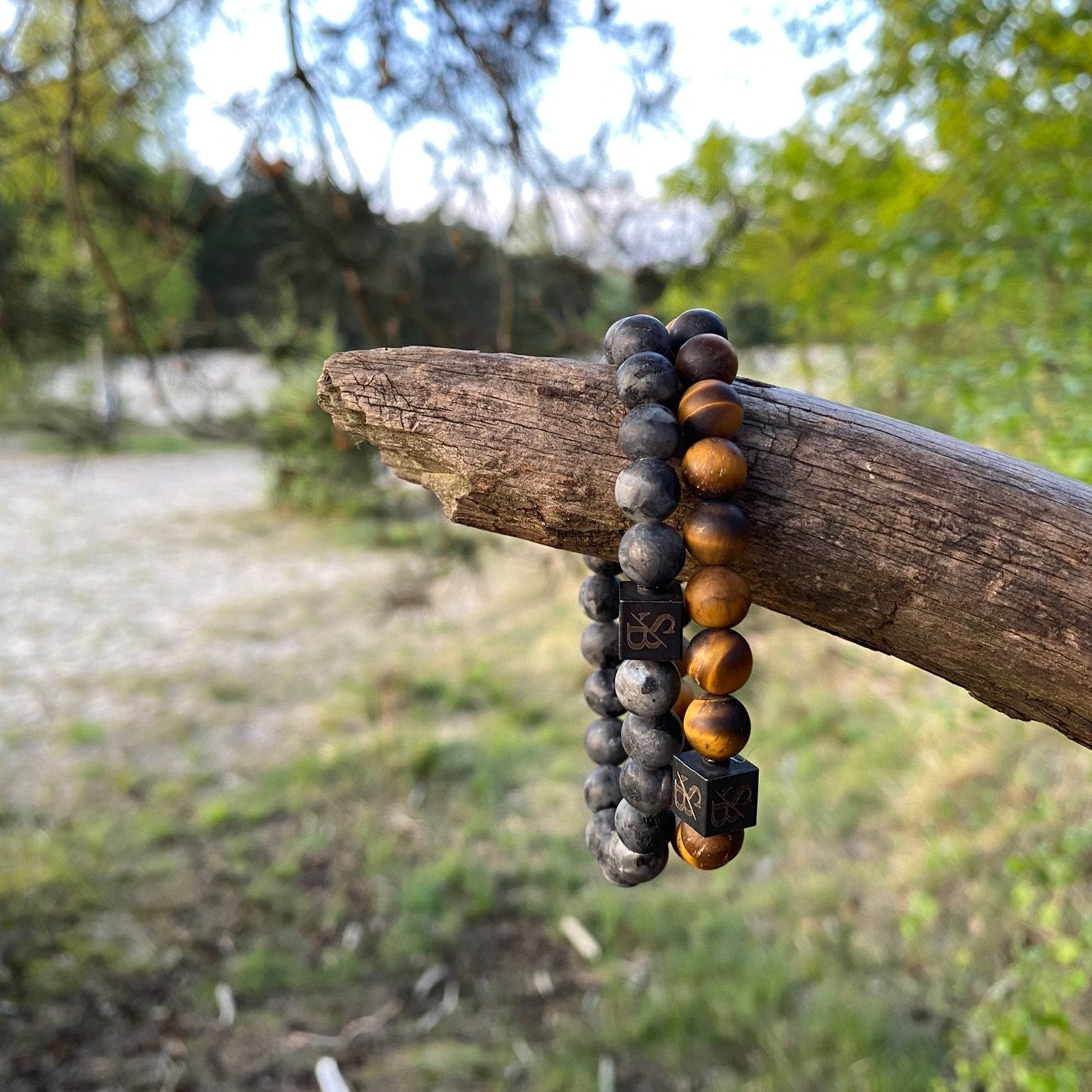 Twee kralenarmbanden van Stoney Bracelets zijn gedrapeerd over een horizontale boomtak buiten. De ene heeft Matte Moon Stones (8 mm) in zwarte en grijze gemarmerde patronen, terwijl de andere bruine en amberkleurige kralen heeft. De achtergrond bestaat uit groen en wazige bomen.