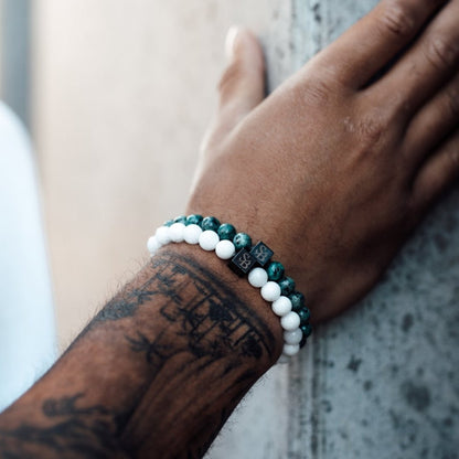 De getatoeëerde onderarm van een persoon rust tegen een muur, versierd met twee kralenarmbanden van Stoney Bracelets. Eén armband heeft 8 mm witte Snowflake Stones, terwijl de andere donkergroene edelstenen heeft.