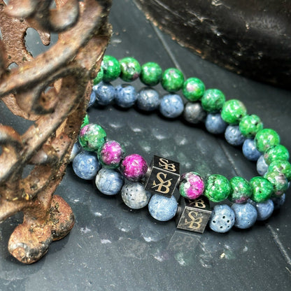 Twee "Coral Stones" armbanden van Stoney Bracelets, gemaakt van 8mm bolvormig natuursteen, zijn gedrapeerd over een decoratief metalen element. De armbanden tonen groene, blauwe en een paar roze gemarmerde kralen, geaccentueerd door kleine vierkante zwarte kralen met een gouden "SB" logo. Het omringende oppervlak en de rekwisieten hebben donkere tinten.