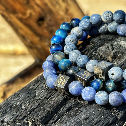 Een close-up van een stapel Stoney Bracelets' Matte Lapis Lazuli Stones (8mm) armbanden, of armbanden, geplaatst op een verkoold, zwartgemaakt stuk hout. De kralen variëren in tinten blauw en omvatten kralen met metalen kubusaccenten met gegraveerde letters. De achtergrond is vervaagd met warme aardse tinten.