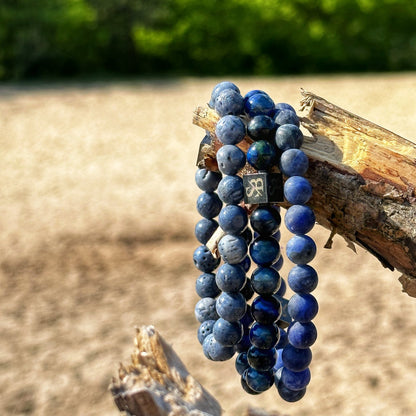 De afbeelding toont de Pacific Set (8mm) van Stoney Bracelets, met drie blauwe kralenarmbanden gemaakt van verschillende tinten edelsteen. Ze zijn gedrapeerd over een gebroken boomtak tegen een wazige achtergrond met wat groen en een zanderig gebied.