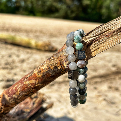 Twee kralenarmbanden uit de Stoney Bracelets Elegance Set (8mm) zijn gedrapeerd over een verweerd stuk drijfhout op een zanderig, zonnig strand met groen op de achtergrond. De door de natuur geïnspireerde armbanden hebben verschillende tinten groene, grijze en witte kralen, geaccentueerd met kleine zwarte blokjes met onduidelijke symbolen.