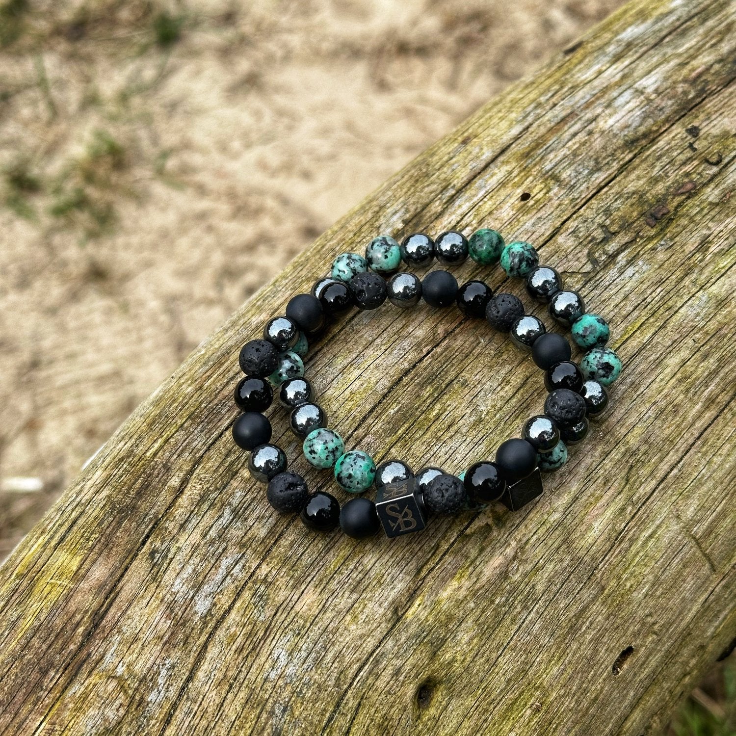 Twee "Prehistoric | Mixed Stones (8mm)" armbanden van Stoney Bracelets zijn op een houten oppervlak geplaatst. De ene heeft een mix van zwarte en groene kralen, terwijl de andere bestaat uit zwarte, grijze en zilverkleurige kralen. Beide armbanden hebben een kleine rechthoekige bedel met een gegraveerd symbool.