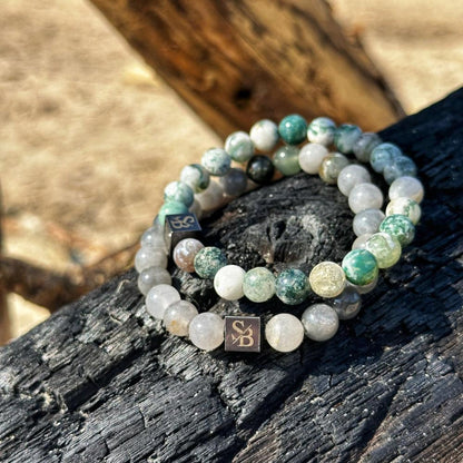 Een paar kralenarmbanden met semi-transparante en groene gemarmerde Tree Agaat Stones (8 mm) van Stoney Bracelets, rustend op een stuk donker, getextureerd hout. Elke armband bevat een kleine metalen kubus gegraveerd met initialen. De achtergrond is wazig.