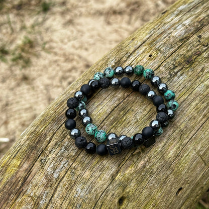 Twee kralenarmbanden van Stoney Bracelets zijn op een verweerd houten oppervlak geplaatst. Eén armband, genaamd "Nautic", is samengesteld uit zwarte kralen, terwijl de andere, genaamd "Mixed Stones (8mm)", een mix van zwarte en groenachtig-turquoise natuursteen kralen bevat. Beide hebben een kleine vierkante bedel met de initialen "SB". De achtergrond is wazig vuil of zand.