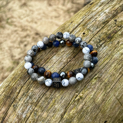 Een set Stoney Bracelets' "Natural | Mixed Stones (8mm)" kralenarmbandarmbanden, met witte, blauwe, grijze en bruine natuursteen, is geplaatst op een verweerd houten oppervlak. De armbanden bevatten een kleine rechthoekige kraal met een gestileerd monogram. Zand is zichtbaar op de achtergrond.