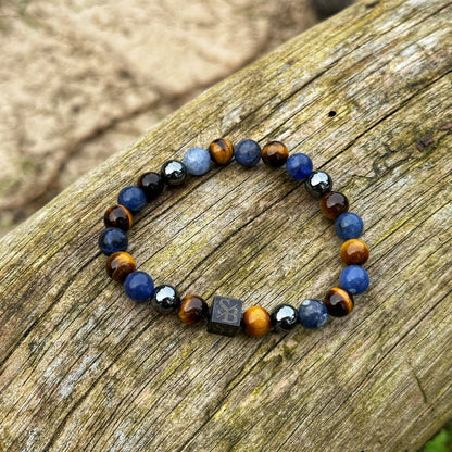 Een Stoney Bracelets Rainforest | Mixed Stones (8mm) armband is geplaatst op een verweerd houten oppervlak. De armband heeft afwisselend blauwe en bruine kralen, aangevuld met een enkele kubieke kraal met een patroon of ontwerp. Het houten oppervlak toont zichtbare nerf en textuur.