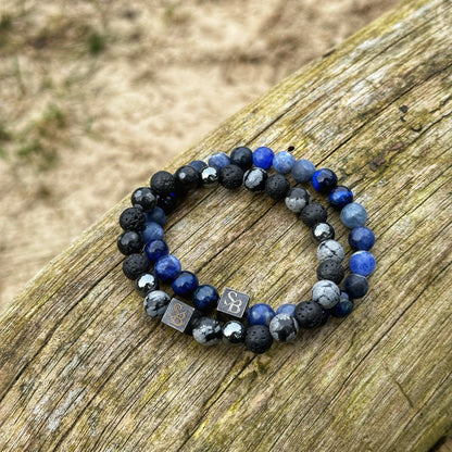 Twee Dark Polar armbanden van Stoney Bracelets, gemaakt met een mix van 8mm blauwe, zwarte en grijze stenen, rusten op een verweerd houten oppervlak. De armbanden zijn versierd met vierkante metalen bedels gegraveerd met de initialen "SB." Zand en gras vormen een wazige achtergrond.