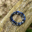 Twee "Ocean | Mixed Stones (8mm)" armbanden van Stoney Bracelets liggen op een houten oppervlak. De armbanden bestaan uit zwarte, blauwe en grijze kralen, waarvan sommige met marmerpatronen en andere met een metallic afwerking. Beide hebben kleine vierkante bedeltjes met een gegraveerd logo, prachtig aangevuld door hun natuurlijke stenen elementen.