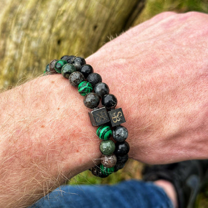 Een pols versierd met twee kralenarmbanden van Stoney Bracelets, met het "Scandinavian | Mixed Stones (8mm)"-ontwerp. Eén armband toont een mix van zwarte en groene natuursteenkralen, terwijl de andere voornamelijk bestaat uit zwarte kralen. Beide stukken zijn geaccentueerd met vierkante metalen bedels gegraveerd met een klavertjevier-ontwerp. De achtergrond benadrukt een houten textuur aangevuld met groen.