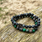 Twee kralen *Volcanic | Mixed Stones (8mm) armbanden* van *Stoney Bracelets*, met ronde zwarte, grijze en groene edelstenen, zijn geplaatst op een verweerd houten oppervlak. Eén armband toont een vierkante zwarte bedel met de inscriptie "SB." De achtergrond bevat een zanderig gebied met hints van gras.