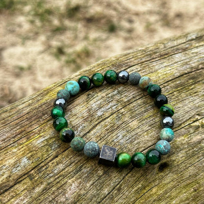 Een armband van Stoney Bracelets, genaamd "Swamp | Mixed Stones (8mm)", met groene, blauwe en zwarte ronde kralen, wordt op een houten oppervlak getoond. Deze elegante armband bevat een opvallende kleine zwarte vierkante kraal met een geëtst ontwerp. De achtergrond is wazig, wat een aardse buitenomgeving laat zien.