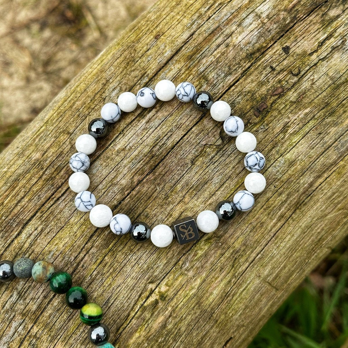 Een Arctic | Mixed Stones (8mm) armband gemaakt van witte en zwarte marmerachtige kralen naast glanzende zwarte kralen ligt op een verweerd houten oppervlak. Er is een kleine vierkante bedel met de initialen "SB" erop, wat staat voor Stoney Bracelets. Een andere groene en zwarte natuursteen kralen armband is gedeeltelijk zichtbaar hieronder.