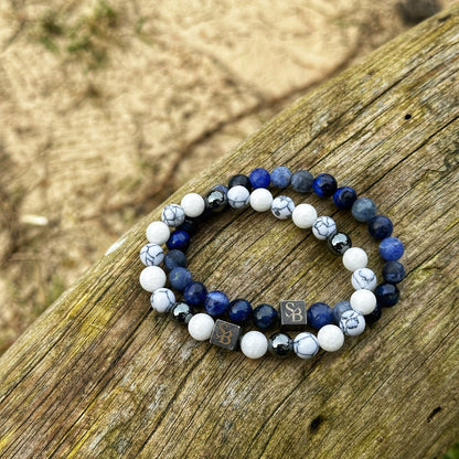 Twee kralenarmbanden uit de Water Set (8mm) van Stoney Bracelets, met een mix van blauwe, witte en zwarte kralen met wat grijs marmer, rusten op een verweerd houten oppervlak. De armbanden zijn versierd met kleine metalen vierkante bedeltjes met de inscriptie "SB". Op de achtergrond is er zand en dun gras.