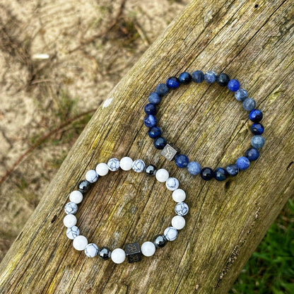 Twee kralen armbanden uit de Stoney Bracelets' "Ocean | Mixed Stones (8mm)" collectie zijn geplaatst op een verweerd houten oppervlak buiten. De bovenste armband is voorzien van donkerblauwe kralen met zilveren accenten, terwijl de onderste armband witte natuursteen kralen met zwarte aders en soortgelijke zilveren accenten toont. De achtergrond bestaat uit gras en zand.