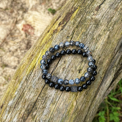 Twee kralenarmbanden zijn op een verweerd houten oppervlak geplaatst. De Dragon Vein Stones (8 mm) van Stoney Bracelets hebben afwisselende patronen van zwarte en gemarmerde grijze kralen, met kleine metalen bedels met in elkaar verstrengelde letters "SB". De achtergrond heeft hints van gras en aarde, wat een vleugje natuursteen aan de scène toevoegt.